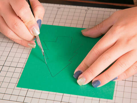 Step1 將美術紙裁剪成合適大小，綠色寬13.5×長14cm、橘紅色寬12.5×長14cm、咖啡色寬15×長14cm。在綠色紙張右側，用鉛筆畫出高度在7公分以內的耶誕樹後，以美工刀切割。（攝影／賴智揚）